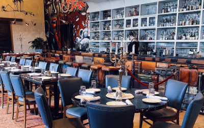 The dining room at fulton market kitchen features blue leather back chairs at a round table with table settings. In the middle sits a silver candelabra. They also have a fully stocked bar to the far right of the restaurant.